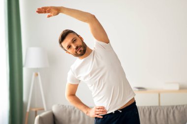 Spor kıyafetli orta yaşlı bir adam evde tek başına egzersiz yapıyor, vücudunu esnetiyor, fotokopi çekiyor. Yakışıklı sakallı adam sabah antrenmanı yapıyor, oturma odasının içinde. Sağlıklı yaşam tarzı kavramı