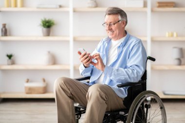 Tekerlekli sandalyedeki yaşlı adam mesajları kontrol ediyor ya da evde akıllı telefondan video izliyor. Mutlu, olgun, engelli erkek cep telefonu kullanıyor, internette geziniyor.