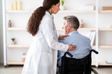 Tekerlekli sandalyedeki yaşlı adam virüs taşıyor, kadın doktor evde nefesini dinliyor. Fiziksel engelli yaşlı hasta covid salgını sırasında genç doktordan tıbbi yardım alıyor.