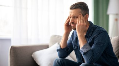 Stresli orta yaşlı sakallı bir adam baş ağrısı çekiyor, evdeki koltukta oturuyor ve şakaklarını ovuyor, fotokopi çekiyor. Migrenden muzdarip üzgün bir adam ya da akşamdan kalma, kafasına dokunuyor.