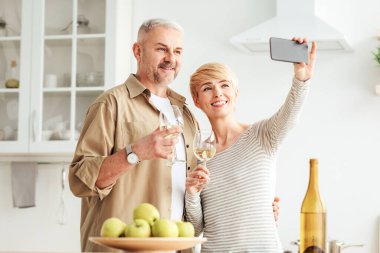 Mutlu romantik çift buluşmanın tadını çıkarıyor ve selfie ya da online görüşme yapıyor. Gülümseyen son sınıf öğrencisi ve şarap kadehli bir kadın minimalist beyaz mutfak içinde meyvelerle akıllı telefondan fotoğraf çekiyorlar.