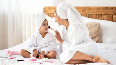 Genç anne gülümseyen tatlı kızına fırçayla dokunuyor, makyaj aletleriyle oynuyor, beyaz bornozlar giyiyor, evde büyük bir yatakta oturuyor, birlikte güzel bir günün tadını çıkarıyor, boşluğu kopyalıyor.