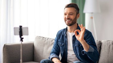 Neşeli orta yaşlı sakallı adam blogcusu akıllı telefon, tripod ve kopyalama alanı kullanarak evden yayın yapıyor. Gülümseyen erkek vlogger cep telefonu kamerasına el sallıyor, takipçiler için video çekiyor