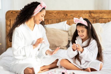 Neşeli anne ve kız evde manikür yapıyor, bornoz giyiyor, yatağa oturuyor ve el tırnaklarına oje sürüyor, güzel bir anne ve küçük bir kız güzel bir gün geçiriyor, yatak odasının içinde.