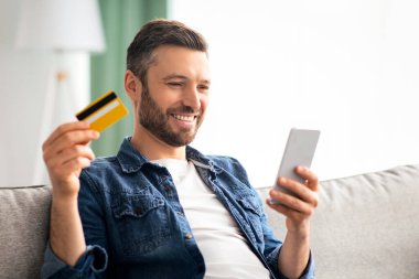 Positive middle-aged man with modern smartphone holding credit card and smiling, shopping online while staying home during COVID-19 pandemic, copy space. E-commerce, online purchasing