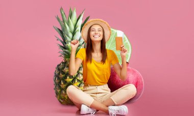 Neşeli bir kadın, geniş meyvelerle çevrili parlak pembe bir arka plana bağdaş kurarak oturur. Bir elinde kredi kartı tutuyor ve geniş bir şekilde gülümsüyor, anın tadını çıkarıyor..