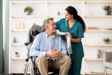 Genç hemşire huzurevinde sıcak ekoseli tekerlekli sandalyedeki yaşlı adamı koruyor. Milenyum bakıcısı engelli yaşlı bir hastaya yardım ediyor, evde yaşlı bir erkeğe bakıyor.