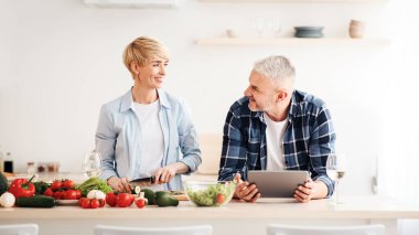 COVID-19 tecridi sırasında romantik bir akşam yemeği için birlikte yemek pişirmek. Gülümseyen yaşlı karı koca konuşuyor, kadın sebzeleri kesiyor, adam tableti minimalist beyaz mutfak içinde tutuyor.