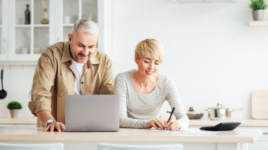 Covid-19 tecrit, fatura ödemesi, ev muhasebesi, yurt içi finans ve vergi. Gülümseyen orta yaşlı beyaz erkek ve kadın, beyaz minimalist mutfak iç dekorasyonunda dizüstü bilgisayarla masrafları göz önünde bulundururlar.