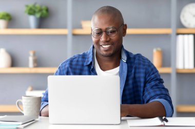 Çevrimiçi kariyer. Dizüstü bilgisayarda yazan yetişkin bir serbest yazar. Bilgisayarda çalışırken gözlük takan Afrikalı Amerikalı bir adam. Ev ofisinin masasında oturuyor. Mesafeli işlerden hoşlanıyor.