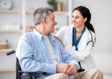 Tekerlekli sandalyedeki yaşlı adam ve onunla evde ilgilenen genç hemşire. Doktorundan profesyonel tıbbi yardım alan mutlu engelli erkek portresi