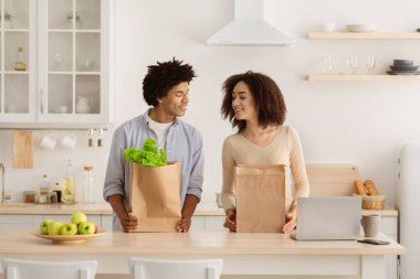 Covid-19 karantinası sırasında marketten teslimat ya da boş vakitlerde alışveriş. Neşeli genç Afro-Amerikan erkek ve dişi. Ekolojik çantalarda yiyecekleri var. Mutfakta birbirlerini izliyorlar.