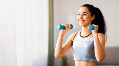 Evde dambıl egzersizi yapan, kol gücü üzerinde çalışan, fotokopi odasına bakan neşeli genç bir kadının yakın çekimi. Gülümseyen atletik kadın oturma odasının iç tarafına mavi fitness damlalarını kaldırıyor.