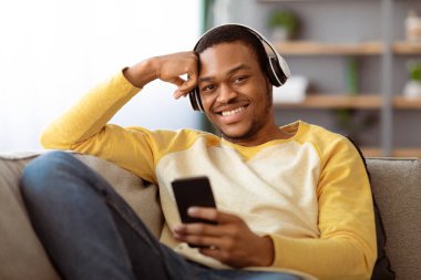 Neşeli siyahi adam evde cep telefonuyla film izliyor, koltuğa yaslanıyor, kablosuz kulaklık kullanıyor, yakın plan çekiyor. Mutlu Afro-Amerikan genç adam akıllı telefondan eğlenceli yeni uygulamalar kullanıyor.