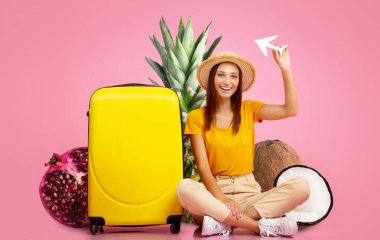 Neşeli bir kadın yerde sarı bir bavulun yanında oturuyor ve ananas ve hindistan cevizi gibi tropikal meyveler. Elinde bir kağıt uçak tutuyor. Seyahat heyecanı ve macerayı simgeliyor..