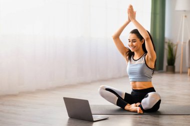 Yoga dersi çevrimiçi. Atletik genç kadın oturma odasında yoga yapıyor, laptopun önündeki yoga minderinde geriniyor, fotokopi çekiyor. Güzel görünümlü esmer kadın evde egzersiz yapıyor, video dersleri alıyor.