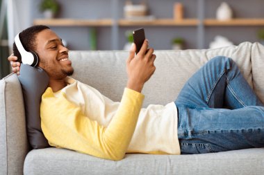 Neşeli Afro-Amerikalı adam evdeki koltukta dinlenirken cep telefonuyla video seyrediyor, kablosuz kulaklık kullanıyor, fotokopi çekiyor. Akıllı telefondan film izleyen neşeli siyahi adam.