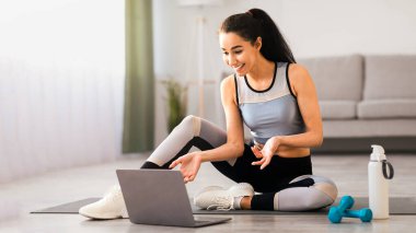 Spor giyim kuşağında gülümseyen güzel bir bayan evden beden eğitimi hocasıyla görüntülü konuşma yapıyor, yoga minderinde halterler ve su şişesiyle oturuyor, dizüstü bilgisayar kullanıyor, konuşuyor ve el kol hareketi yapıyor, boşluğu kopyalıyor.
