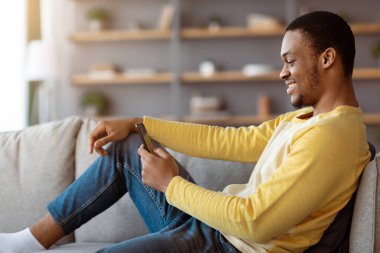 Evdeki kanepede uzanmış gülümseyen siyah adam, akıllı telefon kullanıyor, arkadaşlarıyla sohbet ediyor ya da internette oynuyor, kopya çekiyor. Mutlu Afro-Amerikan genç adam internet sörfü yapıyor veya cep telefonunda sosyal medya kullanıyor.