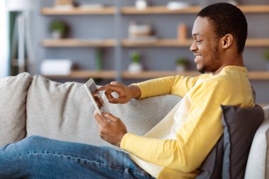 Oturma odasındaki kanepeye yaslanmış, dijital tablet kullanan siyah adamın yan görüntüsü. Mutlu Afro-Amerikan erkeği evde eğleniyor, sosyal medyayı kontrol ediyor ya da arkadaşlarıyla sohbet ediyor.