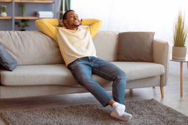 Kaygısız yakışıklı siyahi adam evdeki kanepede dinleniyor, Afro-Amerikan genç adam rahat ve özgür hissederek kanepede uzanırken el ele tutuşuyor ve gülümsüyor.