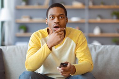 Şok olmuş Afro-Amerikan genç adam evde televizyon kumandası ile oturuyor, haber veya korku filmi izliyor, çenesine, kopyalama alanına, oturma odasına dokunuyor.