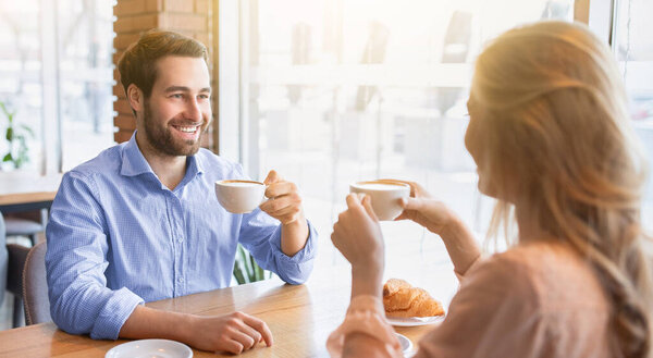 Cheerful young guy having coffee together with his girlfriend at cafe, chatting and smiling during breakfast. Happy millennial couple on romantic date, flirting and spending special moments