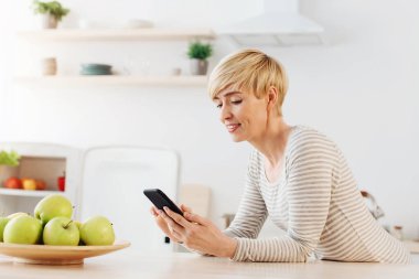 Kahvaltı yaparken cep telefonu, modern cihaz ve internetten teklif, yeni uygulama ve inceleme. Gülümseyen Avrupalı kadın mutfağın içindeki akıllı telefona yazı yazıyor, içi boş.