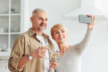 Mutlu romantik çift buluşmanın tadını çıkarıyor ve selfie ya da online görüşme yapıyor. Gülümseyen son sınıf öğrencisi ve şarap kadehli bir kadın minimalist beyaz mutfak içinde meyvelerle akıllı telefondan fotoğraf çekiyorlar.