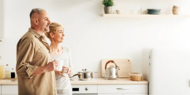 Harika bir teklif, reklam, birlikte kahvaltı, sabah kahvesi, nazik bir ruh hali. Gülümseyen orta yaşlı bir çift sıcak bir bardak içecekle mutfaktaki boş yere bakıyor.