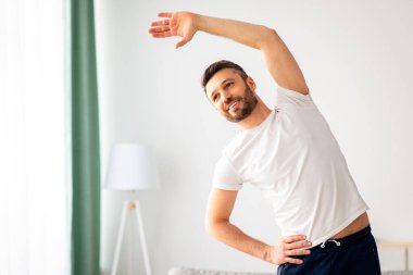 Positif orta yaşlı sakallı bir adam evde spor yapıyor, oturma odasının ortasında duruyor ve vücudunu esnetiyor, sabahları atletizm eğitimi alıyor, uzay kopyaları yapıyor.