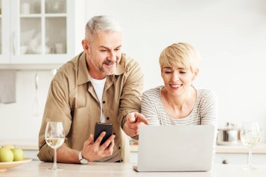 Video görüşmesi, online alışveriş, evdeki cihazlarla çalışma. Telefonu olan mutlu yaşlı bir adam, minimalist mutfakta, içi meyve kadehleriyle dolu, boş bir yerde, olgun bir bayana parmak dizüstü bilgisayarı gösteriyor.