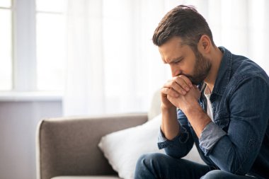 Evdeki kanepede rahat rahat oturan üzgün sakallı, orta yaşlı bir adam. Ellerine yaslanıp yere bakıyor, üzgün bir adamın sorunları var, yan görüş, kopyalama alanı. Yalnızlık, depresyon, akşamdan kalma konsepti.