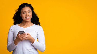Ellerinde akıllı telefon olan genç siyahi kadın ilgiyle fotokopi uzayına bakıyor, neşeli Afrikalı Amerikalı kadın cep telefonu kullanıyor, sarı arka planda tek başına duruyor, Panorama