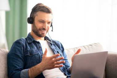 Olumlu sakallı adam evden video konferansı yapıyor, kulaklık ve laptop kullanıyor, konuşuyor ve el kol hareketi yapıyor. Keyifli orta yaşlı bir adam oturma odasındaki koltukta dinleniyor, görüntülü sohbet ediyor, boşluğu kopyalıyor.