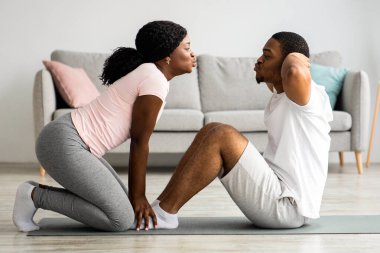 Sportif genç siyahi erkek ve kadın evde birlikte egzersiz yapıyor. Spor kıyafetli Afro-Amerikan kadın erkek arkadaşının bacaklarını tutuyor. Karın kasları üzerinde çalışan adam, yan görüş, kopyalama alanı.