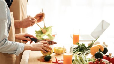 Son yaşlarda sağlıklı beslenme ve vejetaryenlik. Orta yaşlı adam ve önlüklü bayan mutfak içinde salata hazırlayın. Masada sebzeler ve meyve suyu bardakları, kesilmiş, boş alan, kapatın.