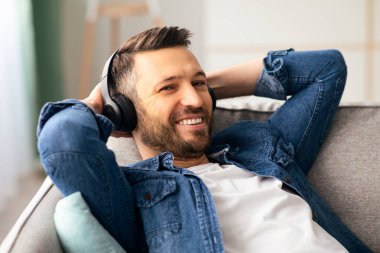 Kablosuz kulaklıkla kanepeye yaslanmış, fotokopi odasına bakıp gülümseyen mutlu sakallı adam. Neşeli orta yaşlı adam evde dinlenirken müzikten zevk alıyor. Müzik dinleme konsepti