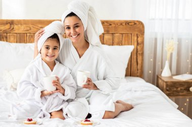 Mutlu genç anne ve kız çay içip yatakta hamur işi yiyorlar, beyaz bornozlar giyip kafalarına havlu geçiriyorlar, evde güzel bir gün geçiriyorlar, uzay kopyaları yapıyorlar.