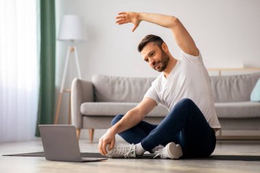 Pozitif atletik sakallı orta yaşlı bir adam evde esniyor, dizüstü bilgisayar kullanıyor, internette spor videoları izliyor, fitness dersi alıyor, kopya çekiyor. Sağlıklı yaşam tarzı, kendini soyutlama sporu.