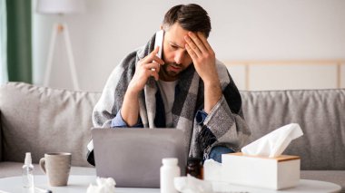 Ateşli orta yaşlı bir adam, doktorunu arıyor ve başına dokunuyor, oturma odasındaki dizüstü bilgisayarın önünde oturuyor, semptomları arıyor ve internetten COVID-19 tedavisi görüyor.