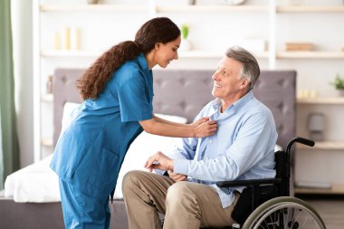 Mutlu genç hemşire, tekerlekli sandalyedeki yaşlı engelli adama yardım ediyor, evde yaşlı bir hastayla ilgileniyor. Milenyum Kafkasyalı bakıcı, olgun engelli erkeğe içeride yardım ediyor.