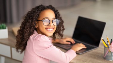 İnternetten ders. Siyah kızın boş ekranlı dizüstü bilgisayarı kullanışının üstünden. Ergen, kameraya bakıyor ve gülümsüyor, evdeki oturma odasında oturuyor.