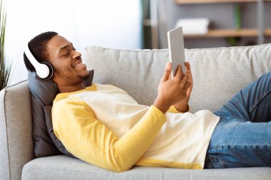 Gülümseyen siyah adam kanepede uzanıyor, evinde film izliyor, fotokopi çekiyor. Mutlu Afro-Amerikan genç adam kanepeye yaslanıyor, modern dijital tablet ve kulaklıklar kullanarak, yan görüş