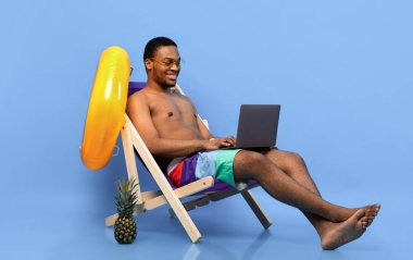 Bilgisayarlı mutlu siyah adam plaj tatilinde çalışıyor, şezlongda oturuyor, bilgisayar klavyesinde yazı yazıyor, mavi stüdyo geçmişi var. Afrika kökenli Amerikalı adam yaz tatillerinde internet belgelerini kontrol ediyor