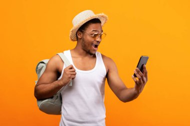 Sırt çantalı havalı siyahi adam akıllı telefonuna heyecanla bakıyor internetten son dakika seyahat turu ayarlıyor, sarı stüdyo geçmişi var. Afrikalı Amerikalı adam büyük indirim ve indirimi öğreniyor.