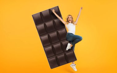 Uzun saçlı bir kadın heyecandan zıplıyor. Elinde kocaman bir çikolatayla parlak sarı bir zemin tutuyor. Bu eğlenceli anın tadını çıkarırken ifadesi saf mutluluğu yansıtıyor..