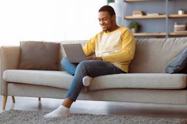 Siyahi adam evde vakit geçiriyor, modern dizüstü bilgisayar kullanıyor, koltukta oturuyor, internette sörf yapıyor, fotokopi çekiyor. Gülümseyen Afrikalı Amerikalı genç adam, bilgisayar klavyesinde daktilo çalıyor, internette sohbet ediyor.