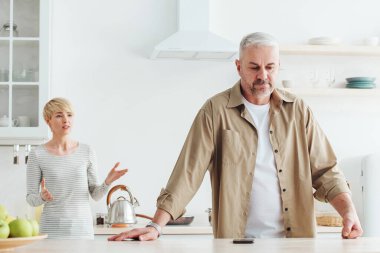 Öfkeli eş kocasını suçluyor, evde evlilik sorunları var. Küfür eden olgun, beyaz adam mutfak içinde küfreden kızgın kadını dinliyor, panorama.