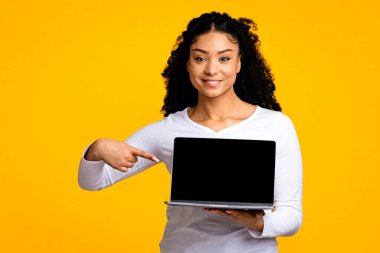 Elinde siyah ekranla dizüstü bilgisayarı işaret eden gülümseyen Afrikalı Amerikalı kadın, web sitesi tasarımı için kopyalama alanı belirten neşeli siyahi kadın, sarı arka planda tek başına duruyor,